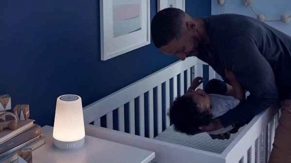 Người cha nhẹ nhàng đặt em bé vào cũi trong phòng bé, với đèn ngủ sáng mờ và bức tranh treo tường nhã nhặn.