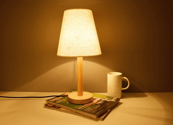 Đèn bàn với ánh sáng ấm áp đặt trên tạp chí cạnh cốc sứ trắng, tạo không gian thư giãn và gần gũi.