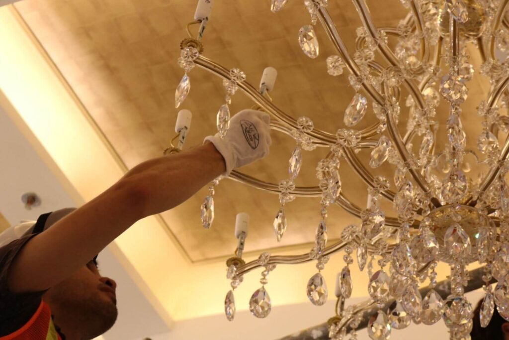 Người thợ đeo găng tay trắng đang lau đèn chùm pha lê lộng lẫy, tạo ra không gian sang trọng và sáng lấp lánh.